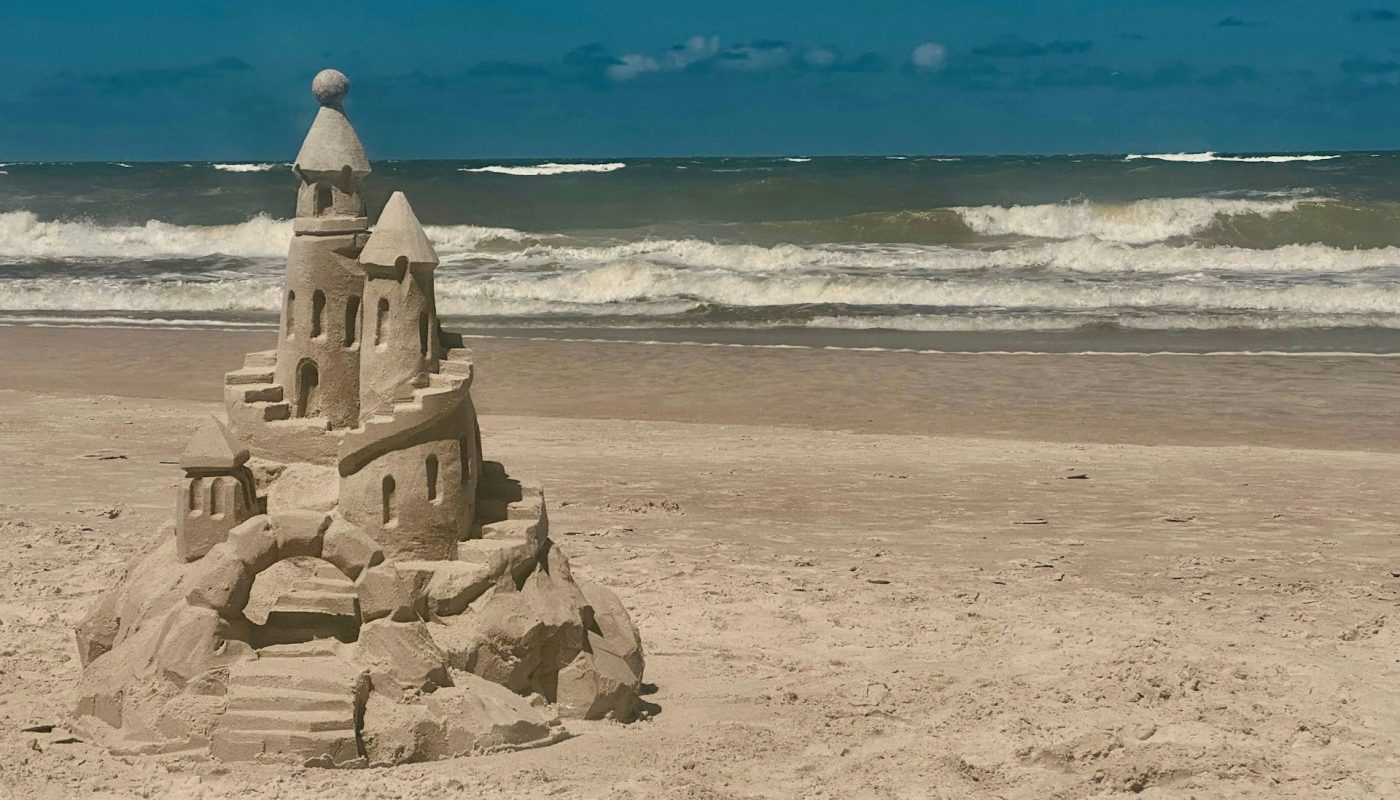 A single sand castle on an open beach, its base already crumbling, with waves rolling in behind it