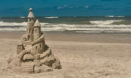 A single sand castle on an open beach, its base already crumbling, with waves rolling in behind it